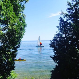 A gap in the trees showing a sailboat and kayak on the lake passing by.